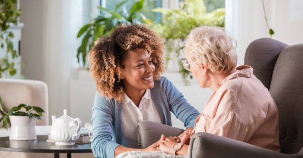 Female home caregiver talking with senior woman, sitting in living room and listening to her carefully.